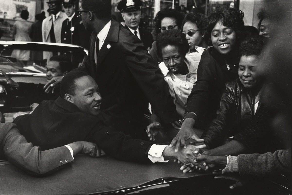 A photo of Martin Luther King Jr. shaking hands with numerous supporters from the back seat of a car.
