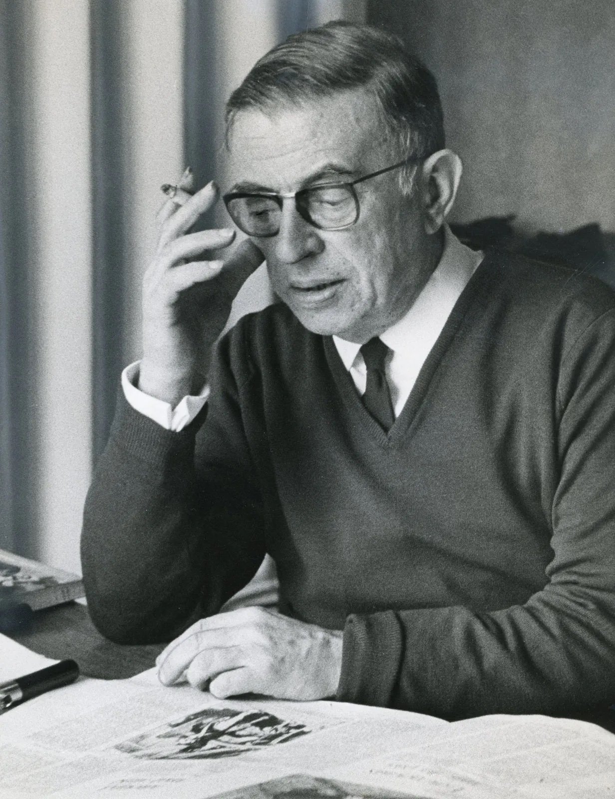 A photograph of Jean Paul Sartre sitting behind a desk, smoking a cigarette and reading a newspaper